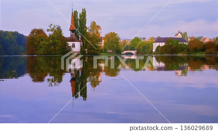 Scenic view of a church and houses by the water reflecting at sunrise in a quiet town Scenic view of a church and houses by the water reflecting at sunrise in a quiet town 136029689