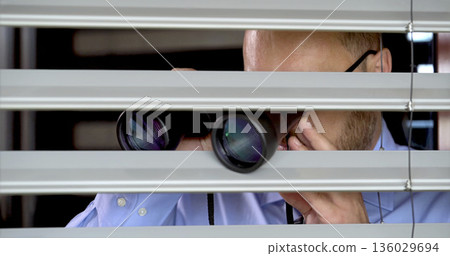 Man looks through blinds with binoculars in an office setting during daylight hours Man looks through blinds with binoculars in an office setting during daylight hours 136029694
