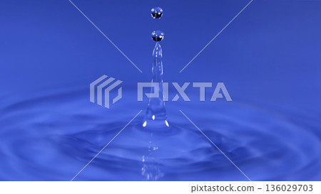 Water drop creates splash in blue background during a close up view at daytime in a simple setting Water drop creates splash in blue background during a close up view at daytime in a simple setting 136029703