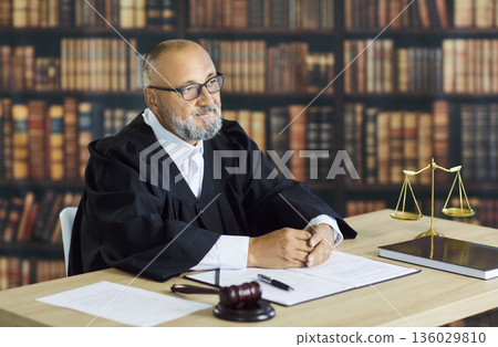 Experienced judge in robe at desk with gavel and scales, in grand law library 136029810