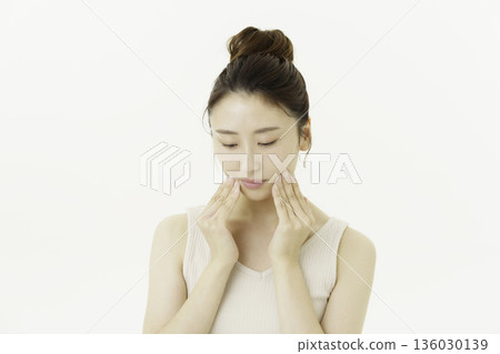 A young Japanese woman wearing a tank top with updo and looking down and touching her cheek against a white background 136030139