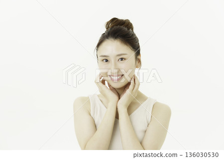 A young Japanese woman wearing a tank top against a white background, with her hair up and smiling with her hand on her cheek 136030530
