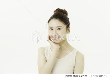 A young Japanese woman wearing a tank top against a white background, with her hair up and smiling with her hand on her cheek 136030532