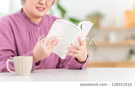 An Asian senior woman sitting at a table and reading. 136031010
