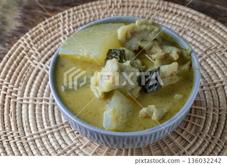 Homemade green curry featuring white gourd and fish balls is served in a gray ceramic bowl set on a circular woven placemat. 136032242