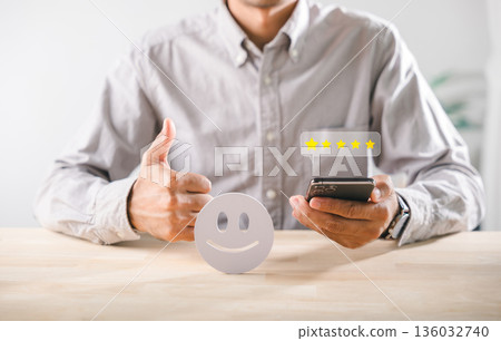Positive customer feedback evaluation showing client satisfaction, 5 star rating on smartphone, happy service experience with person giving thumbs up gesture 136032740