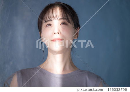 A woman posing in the sunlight against a light blue background. Her upper body has a dazzling expression. She is wearing sunscreen and UV protection. 136032796