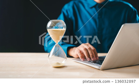 Hourglass showing time passing, sand glass clock concept highlighting business urgency for project growth, person diligently works on laptop for deadline countdown, optimizing valuable hour 136032897