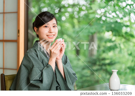 A young Asian woman in a yukata haori relaxing on the veranda of a guest room in a ryokan, drinking sake A young Asian woman in a yukata haori relaxing on the veranda of a guest room in a ryokan, drinking sake 136033209