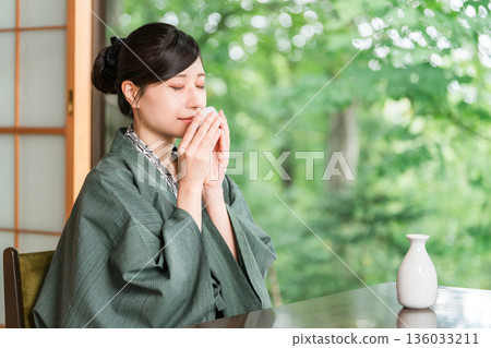 A young Asian woman in a yukata haori relaxing on the veranda of a guest room in a ryokan, drinking sake 136033211