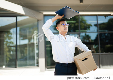 Businessman holding box and file after being fired, showing sadness and uncertainty while leaving modern corporate office building. Businessman holding box and file after being fired, showing sadness and uncertainty while leaving modern corporate office building. 136033431