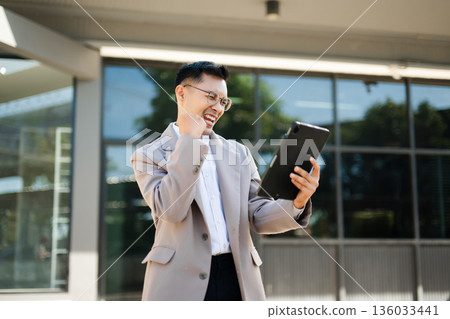 Happy businessman raising fist and cheering with smartphone in hand, celebrating success and victory with excitement and joy outdoors. Happy businessman raising fist and cheering with smartphone in hand, celebrating success and victory with excitement and joy outdoors. 136033441