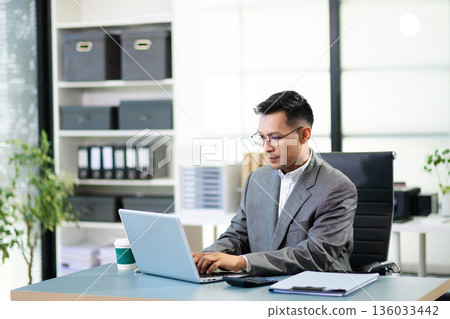Asian man using laptop and tablet while sitting at her working place. Concentrated at work. Asian man using laptop and tablet while sitting at her working place. Concentrated at work. 136033442