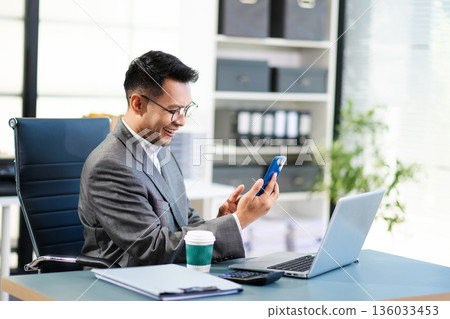 Asian man using laptop and tablet while sitting at her working place. Concentrated at work. Asian man using laptop and tablet while sitting at her working place. Concentrated at work. 136033453