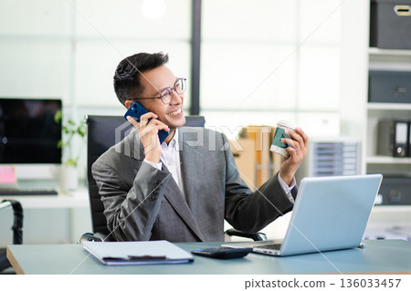 Businessman managing multiple tasks, talking on phone, checking reports, and working on laptop in a busy modern corporate office. Businessman managing multiple tasks, talking on phone, checking reports, and working on laptop in a busy modern corporate office. 136033457