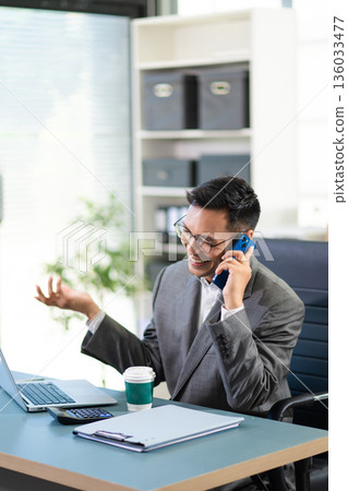 Businessman managing multiple tasks, talking on phone, checking reports, and working on laptop in a busy modern corporate office. 136033477