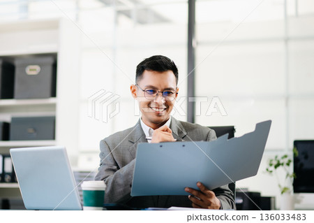 A professional businessman working on a laptop while holding a coffee cup in a modern office, representing productivity A professional businessman working on a laptop while holding a coffee cup in a modern office, representing productivity 136033483