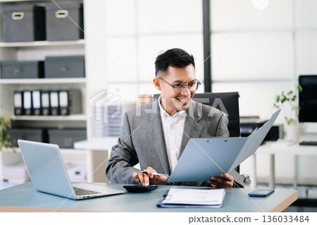 A professional businessman working on a laptop while holding a coffee cup in a modern office, representing productivity 136033484