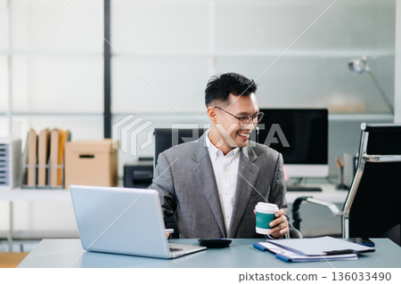 A professional businessman working on a laptop while holding a coffee cup in a modern office, representing productivity 136033490
