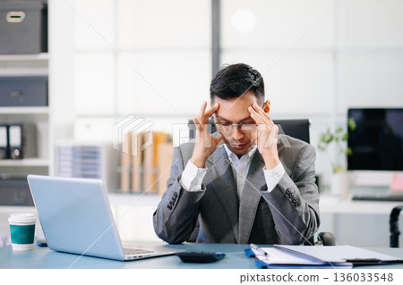 Exhausted businessman sitting at desk with laptop, stressed by workload, deadlines, and corporate pressure in modern office environment. Exhausted businessman sitting at desk with laptop, stressed by workload, deadlines, and corporate pressure in modern office environment. 136033548