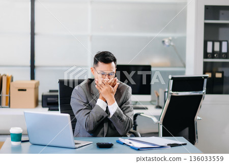 Tired businessman sitting at office desk with laptop, feeling stressed and overworked under tight deadlines 136033559