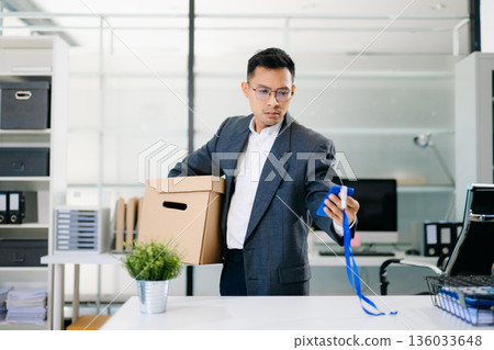 Depressed businessman packing box after job loss, feeling sad and stressed from layoff or resignation in modern office environment. Depressed businessman packing box after job loss, feeling sad and stressed from layoff or resignation in modern office environment. 136033648