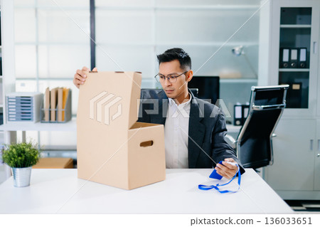 Depressed businessman packing box after job loss, feeling sad and stressed from layoff or resignation in modern office environment. Depressed businessman packing box after job loss, feeling sad and stressed from layoff or resignation in modern office environment. 136033651