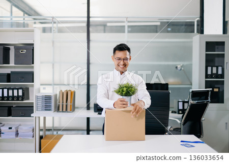 Asian office worker packing belongings into a cardboard box, representing resignation, job change, or a fresh career start in a modern workplace environment. Asian office worker packing belongings into a cardboard box, representing resignation, job change, or a fresh career start in a modern workplace environment. 136033654