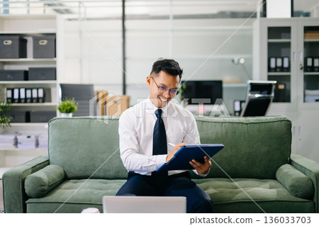 Asian businessman working with a tablet in a modern office, smiling confidently while reviewing documents. Asian businessman working with a tablet in a modern office, smiling confidently while reviewing documents. 136033703