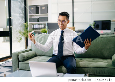 Asian businessman sitting on office sofa using smartphone and laptop, smiling while working with documents in a modern workspace. 136033718