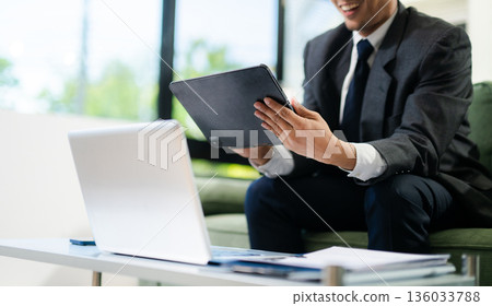Smiling businessman using laptop and smartphone while working in modern office, digital communication, mobile technology, productivity 136033788