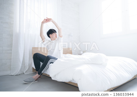 A young man waking up in bed bathed in the morning sun stretching his arms A young man waking up in bed bathed in the morning sun stretching his arms 136033944