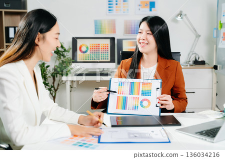 Two creative female designers discussing color palettes and UX UI project in a modern office. Perfect for teamwork, innovation, and digital design themes. 136034216