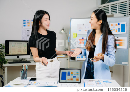 Two UX UI designers collaborating on mobile app wireframes and layouts in modern office. Concept of creativity, teamwork, and digital product design. 136034226