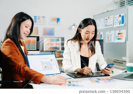Two professional women designers discussing UX UI and color palette in a creative modern office. Ideal for teamwork, innovation 136034300