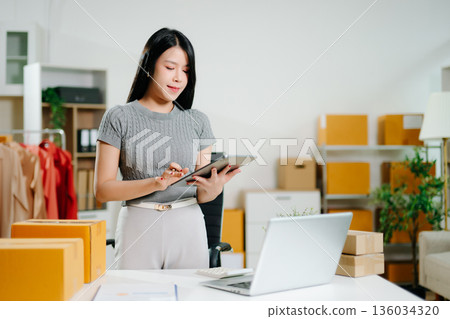 Startup small business SME, Entrepreneur owner woman using smartphone or tablet taking receive and checking online purchase shopping order to preparing pack product box. 136034320