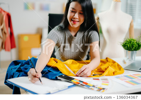 Asian fashion designer measuring fabric at home studio with mannequin and laptop. Creative fashion business, tailor planning clothing design Asian fashion designer measuring fabric at home studio with mannequin and laptop. Creative fashion business, tailor planning clothing design 136034367
