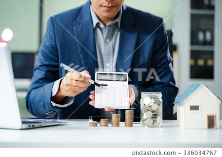 Asian businessman stacking coins beside a house model and jar of savings, planning financial growth and investment strategy. Ideal for finance, banking Asian businessman stacking coins beside a house model and jar of savings, planning financial growth and investment strategy. Ideal for finance, banking 136034767