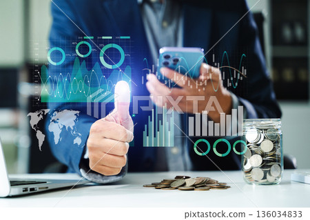 Businessman uses smartphone for virtual fintech analysis. Coins, charts, and futuristic UI symbolize financial success, investments Businessman uses smartphone for virtual fintech analysis. Coins, charts, and futuristic UI symbolize financial success, investments 136034833
