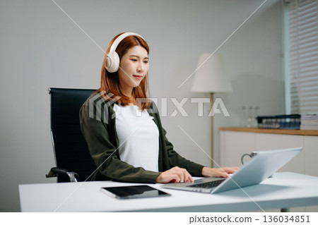Young Asian woman using wireless headphones while typing on laptop in a modern office. 136034851