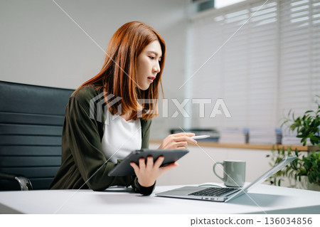 Confident Asian businesswoman working with digital tablet and laptop in bright office. Confident Asian businesswoman working with digital tablet and laptop in bright office. 136034856