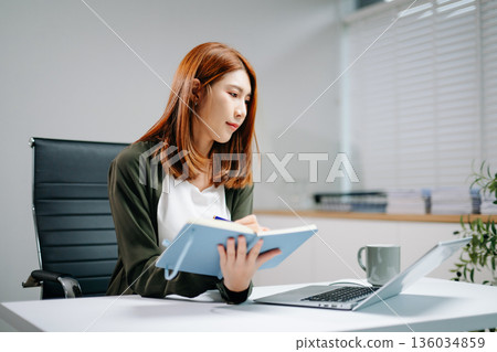 Young Asian woman in office focusing on laptop and writing in notebook. Concept of productivity, online learning, work Young Asian woman in office focusing on laptop and writing in notebook. Concept of productivity, online learning, work 136034859