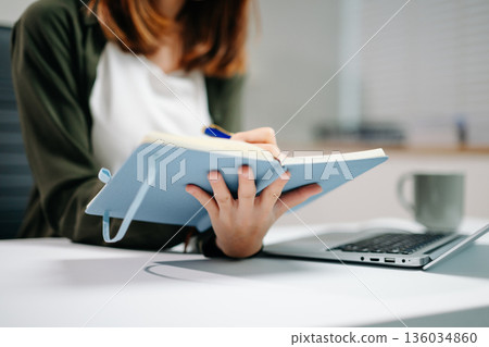 Young Asian woman in office focusing on laptop and writing in notebook. Concept of productivity, online learning, work Young Asian woman in office focusing on laptop and writing in notebook. Concept of productivity, online learning, work 136034860
