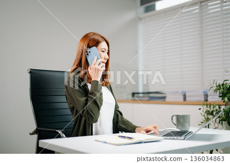 Smiling Asian woman speaks on phone while working at desk with laptop and documents. Concept of communication, teamwork, remote work, and professional success. 136034863