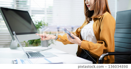 Confident Asian business people using laptops, tablets, and phones in modern office workspace, planning data strategy and success. Confident Asian business people using laptops, tablets, and phones in modern office workspace, planning data strategy and success. 136034898