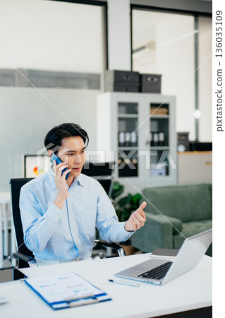 Asian Businessman Talking on the phone and using a laptop with a smile while sitting at office 136035109