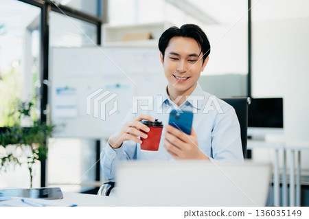 Confident  Asian businessman typing laptop computer and digital tablet while holding coffee at office 136035149