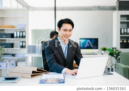 Confident Asian businessman in suit working on laptop and clipboard at bright modern office, symbolizing professionalism, success, and corporate teamwork. 136035161