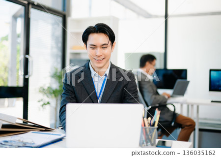 Confident Asian businessman in suit working on laptop and clipboard at bright modern office, symbolizing professionalism, success, and corporate teamwork. 136035162