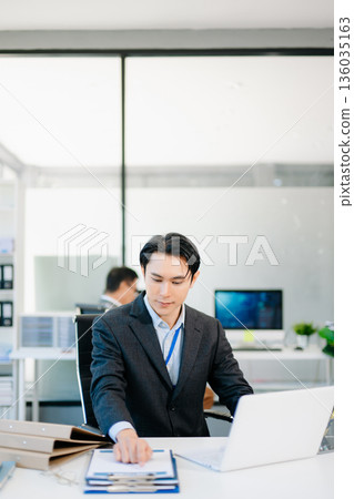 Confident Asian businessman in suit working on laptop and clipboard at bright modern office, symbolizing professionalism, success, and corporate teamwork. 136035163